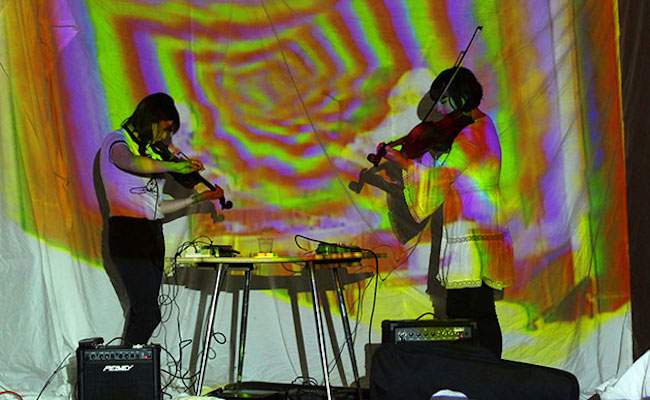 Two people playing the violin with a projected Tie Dye background