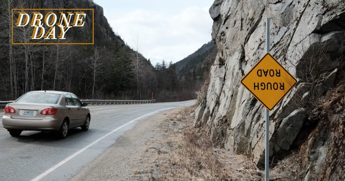 A car driving on a road on a mountain with an upside down road sign that says 'ROUGH ROAD'